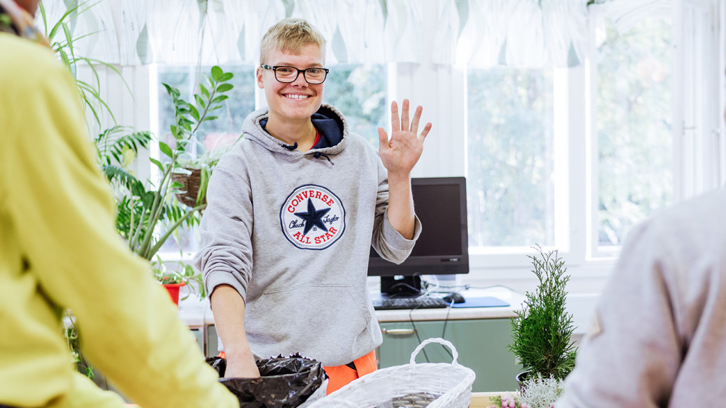 Nuorella on toinen käsi multapussissa ja toisella kädellä hän moikkaa iloisesti.