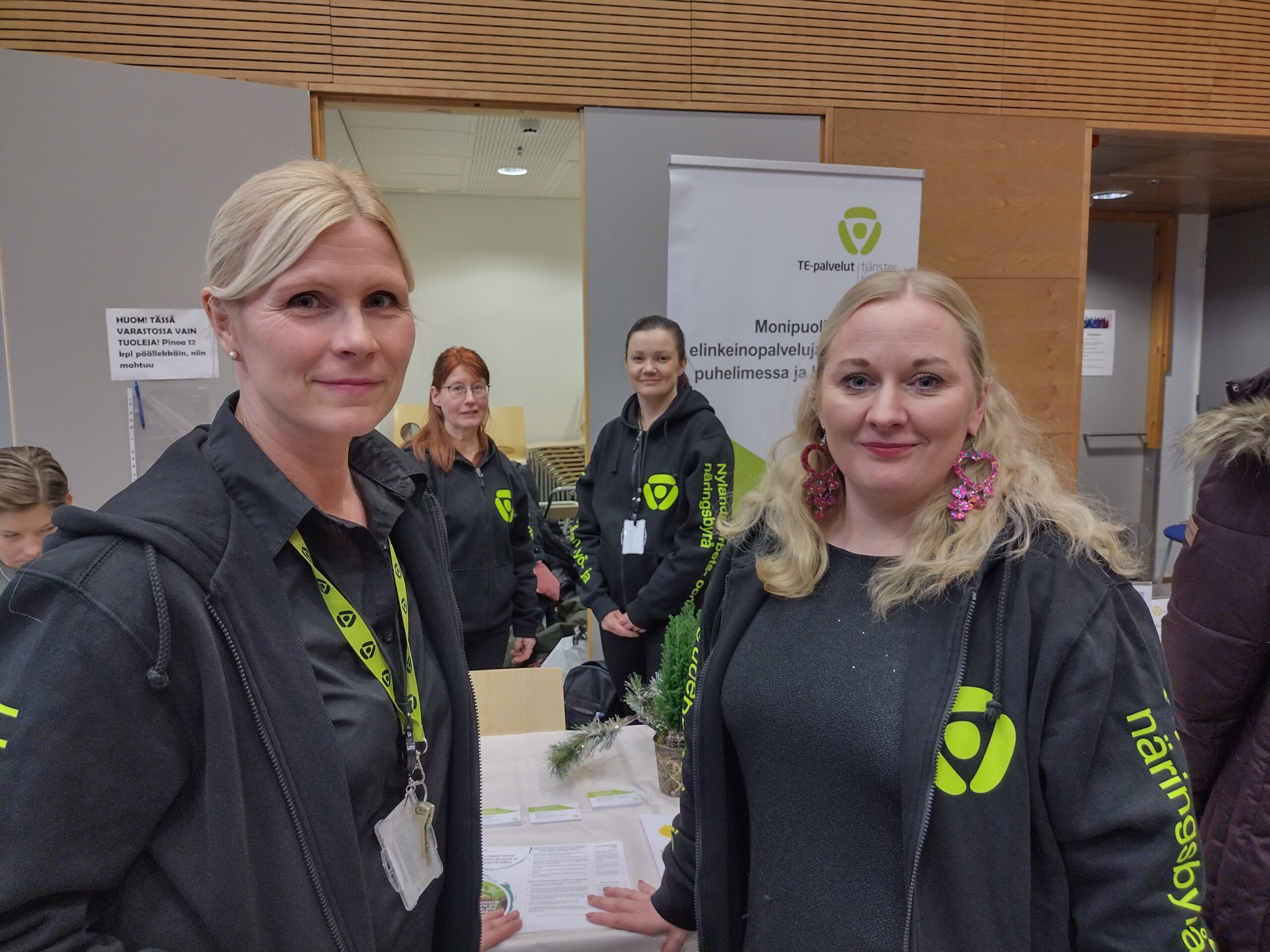 Neljä naista katsoo kameraan tapahtumatilassa, yllään heillä kaikilla on TE-toimiston logolla varustetut mustat hupparit.
