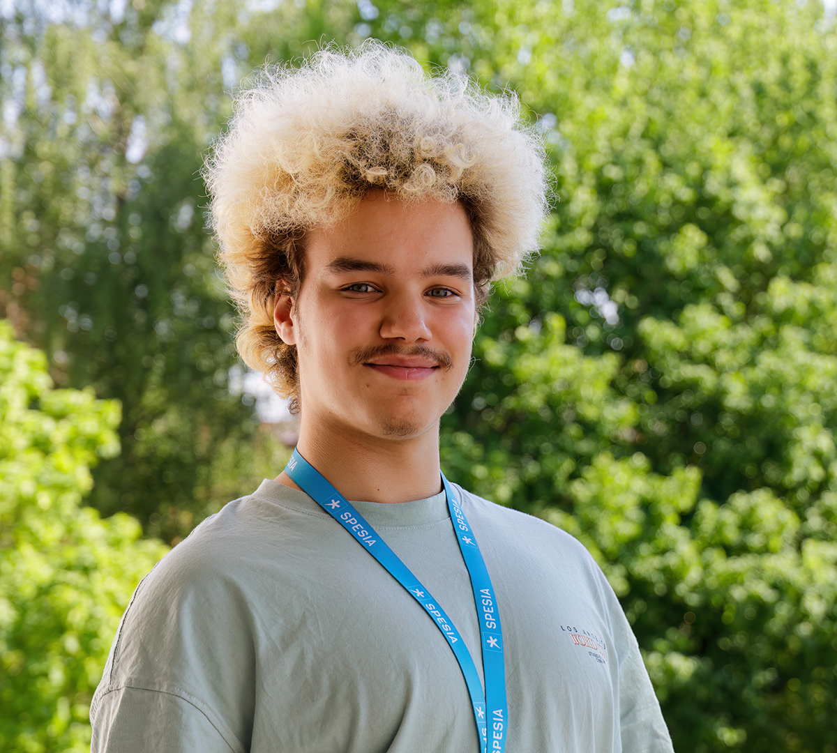 Kiharatukkainen nuori mies t-paidassa ja kaulassa Spesian kaulanauha, taustalla kesäistä näkymää.