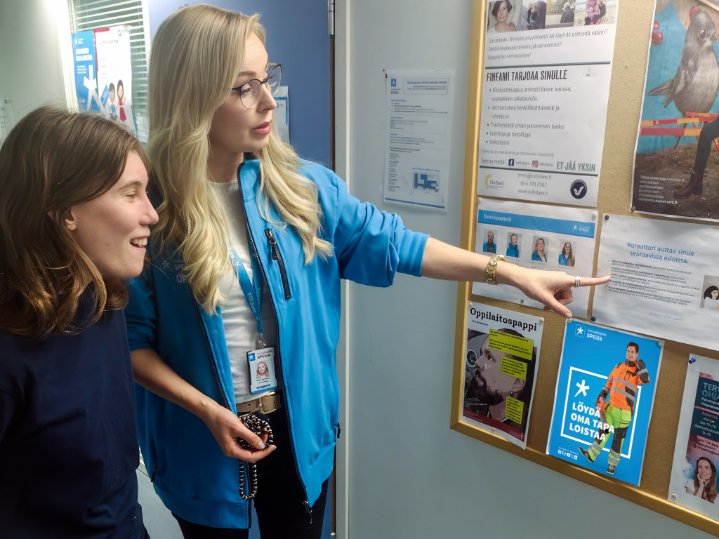 Spesian huppariin pukeutunut aikuinen osoittaa nuorelle ilmoitustaulua.