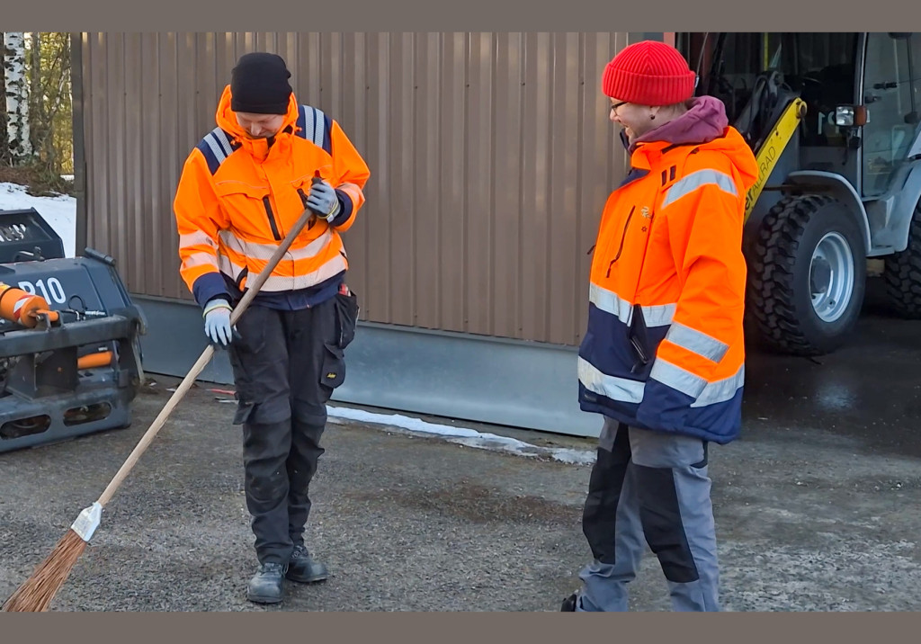 Kaksi miestä oransseissa työtakeissa: toinen lakaisee pihaa ja toinen seuraa tyytyväisen näköisenä työn sujumista.