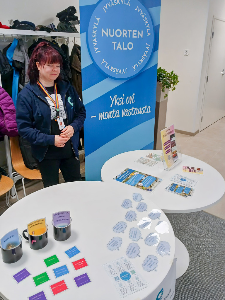 Nainen seisoo esittelypisteella, taustalla Nuorten talon roll-up ja pöydällä kortteja.