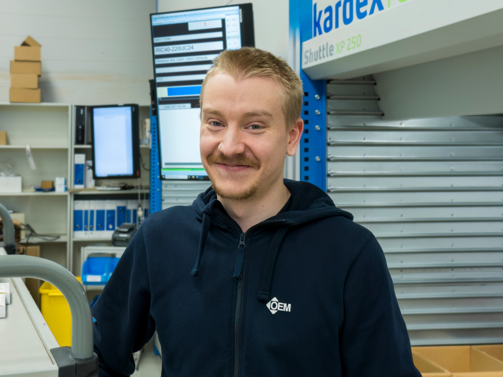 Mies katsoo kameraan hymyillen, taustalla näkyy logistiikkakeskuksen toimitilaa.