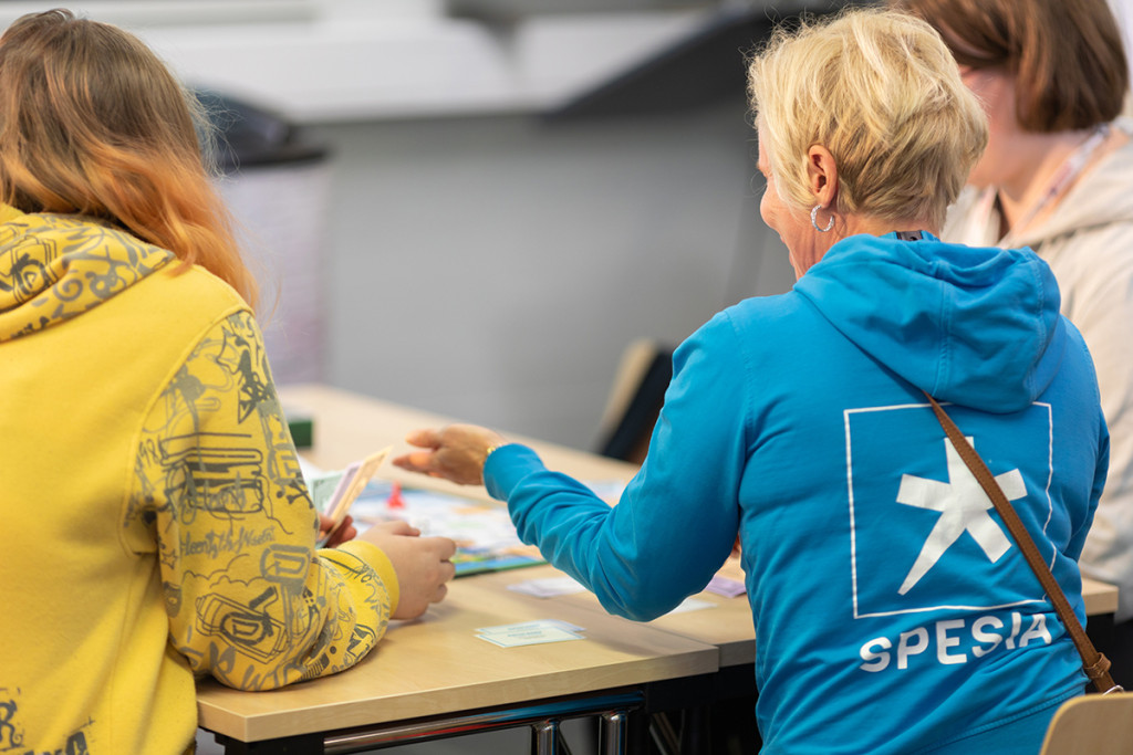 Pöydän ääressä kaksi henkilöä selkä katsojaan päin, toisella Spesian huppari päällä ja toinen näyttää opiskelijalta.