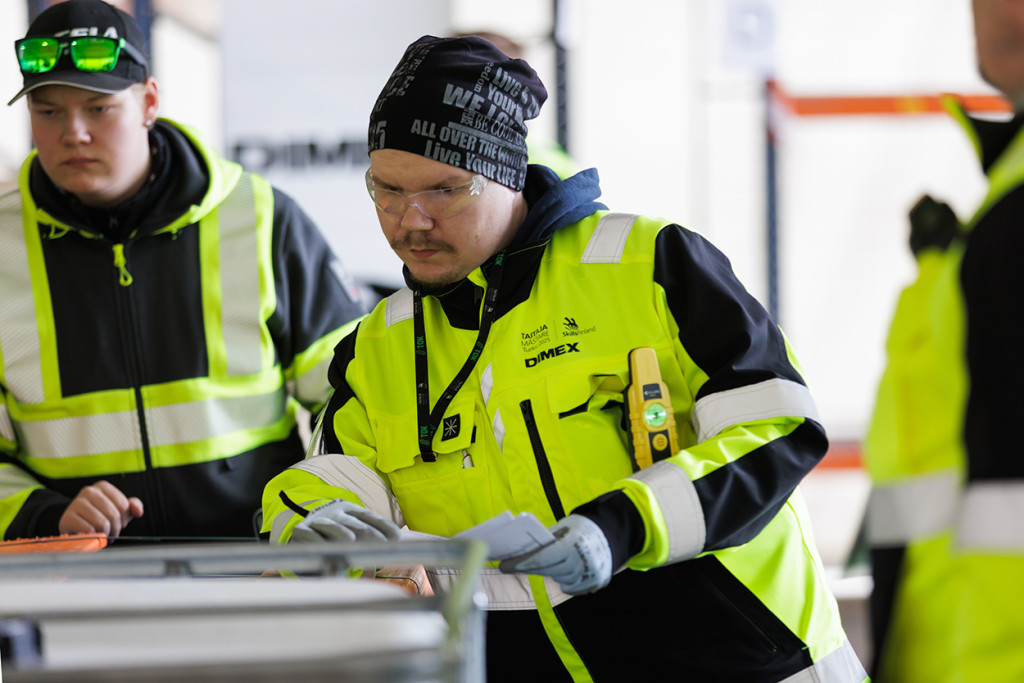 Kelta-mustiin työvaatteisiin pukeutunut Taitaja-kilpailija katsoo tarkasti jotain, kaksi tuomaria seuraa vierestä.
