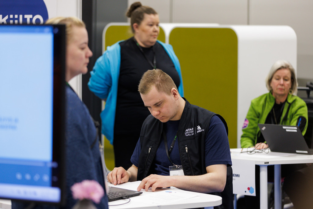 Nuori mies työskentelee tietokoneella Taitaja-kisoissa, kun ympärillä seisoo kolme kisatuomaria.