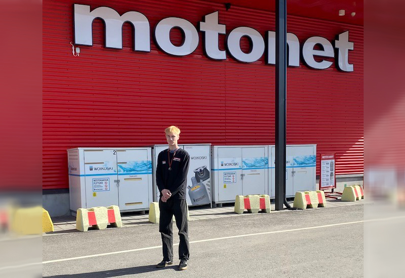 Nuori mies seisoo Motonetin edessä tumma työasu päällä.