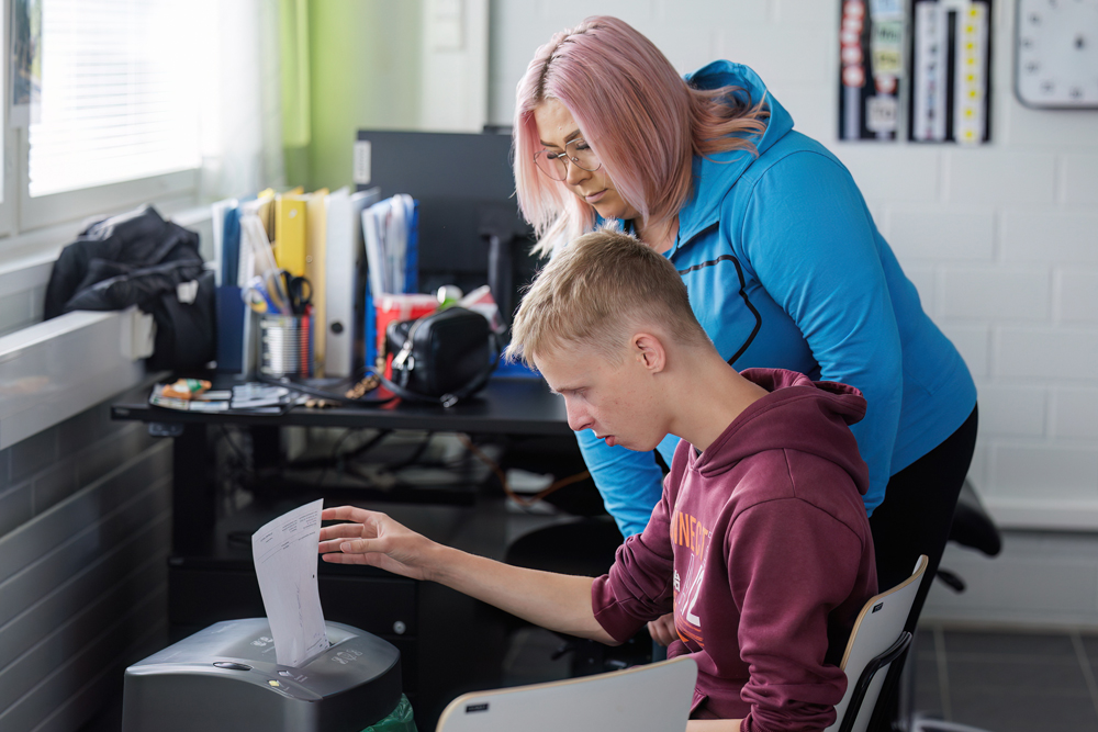 Nuori syöttää paperiarkkia silppuriin, Spesian huppariin pukeutunut henkilö seuraa vierestä.