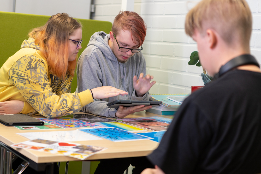 Kolme nuorta opiskelijaa pöydän ympärillä, kaksi tutkii tablettia, pöydällä kuvia ja papereita.