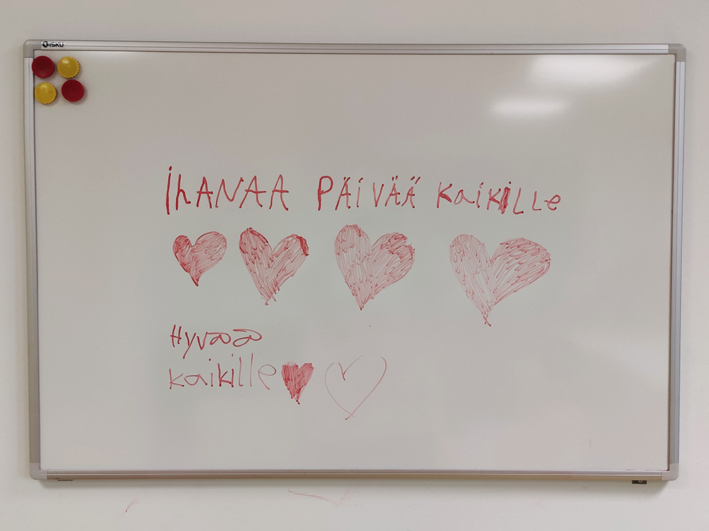 Valkotaululla teksi "Ihanaa päivää kaikille" ja sydämiä.