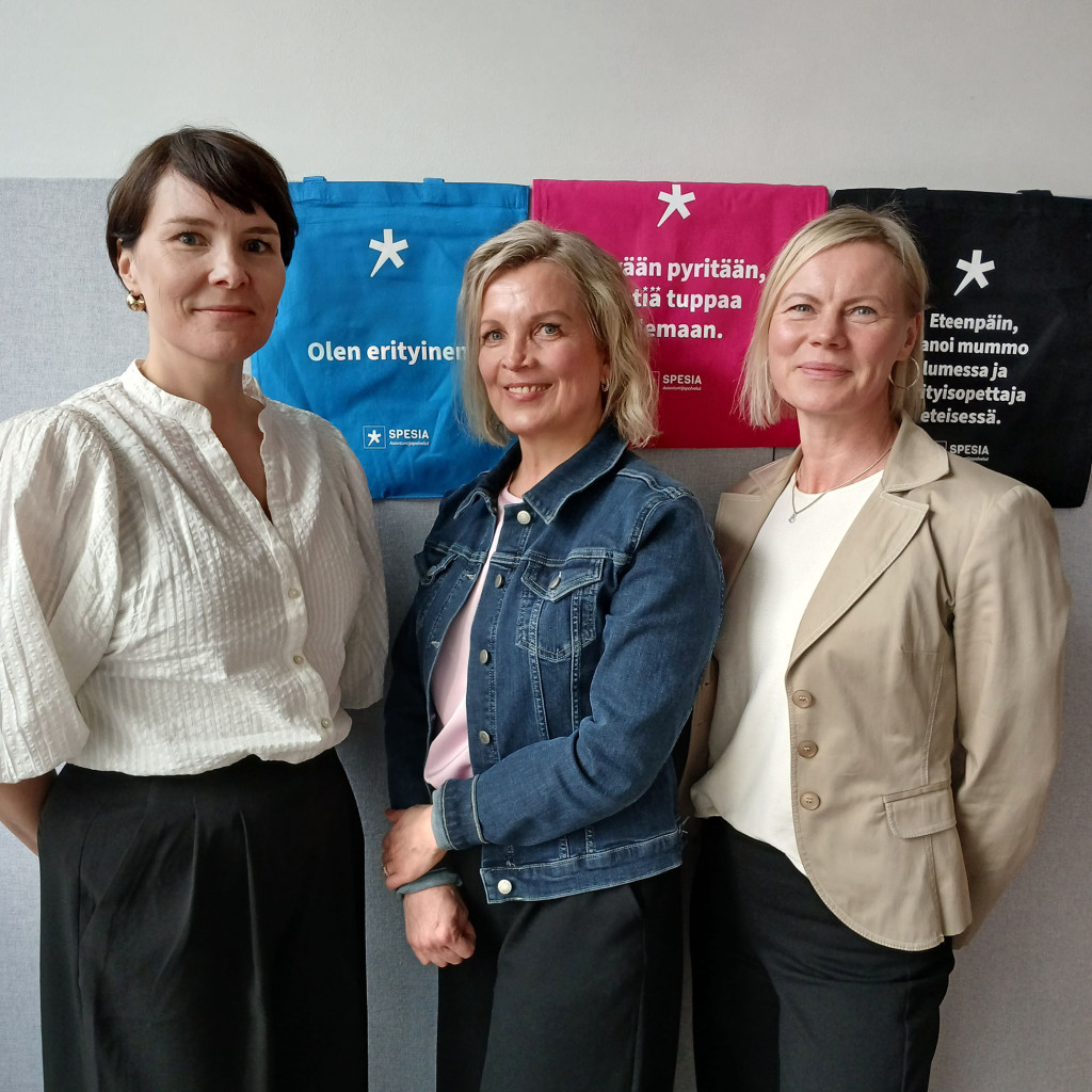 Kolme naista poseeraa, taustalla sininen, pinkki ja musta Spesian kangaskassi.