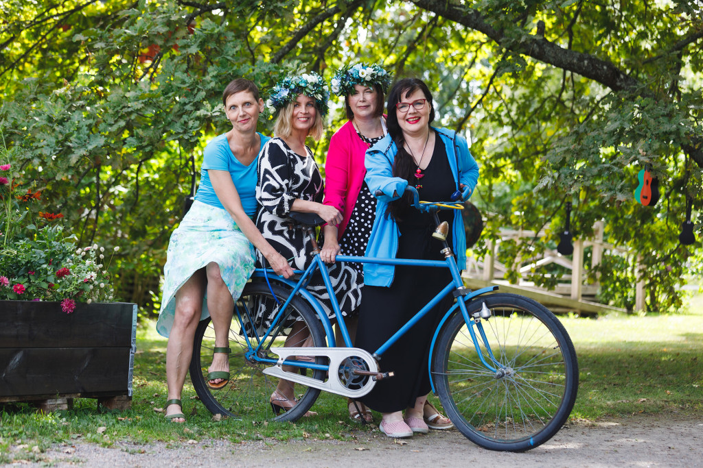 Neljä naishenkilöä ulkona vanhan sinisen polkupyörän kanssa.
