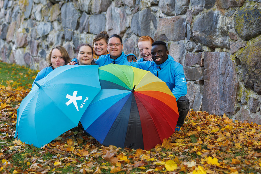 Kuusi henkilöä kurkkii kahden ison maassa olevan sateenvarjon takaa.