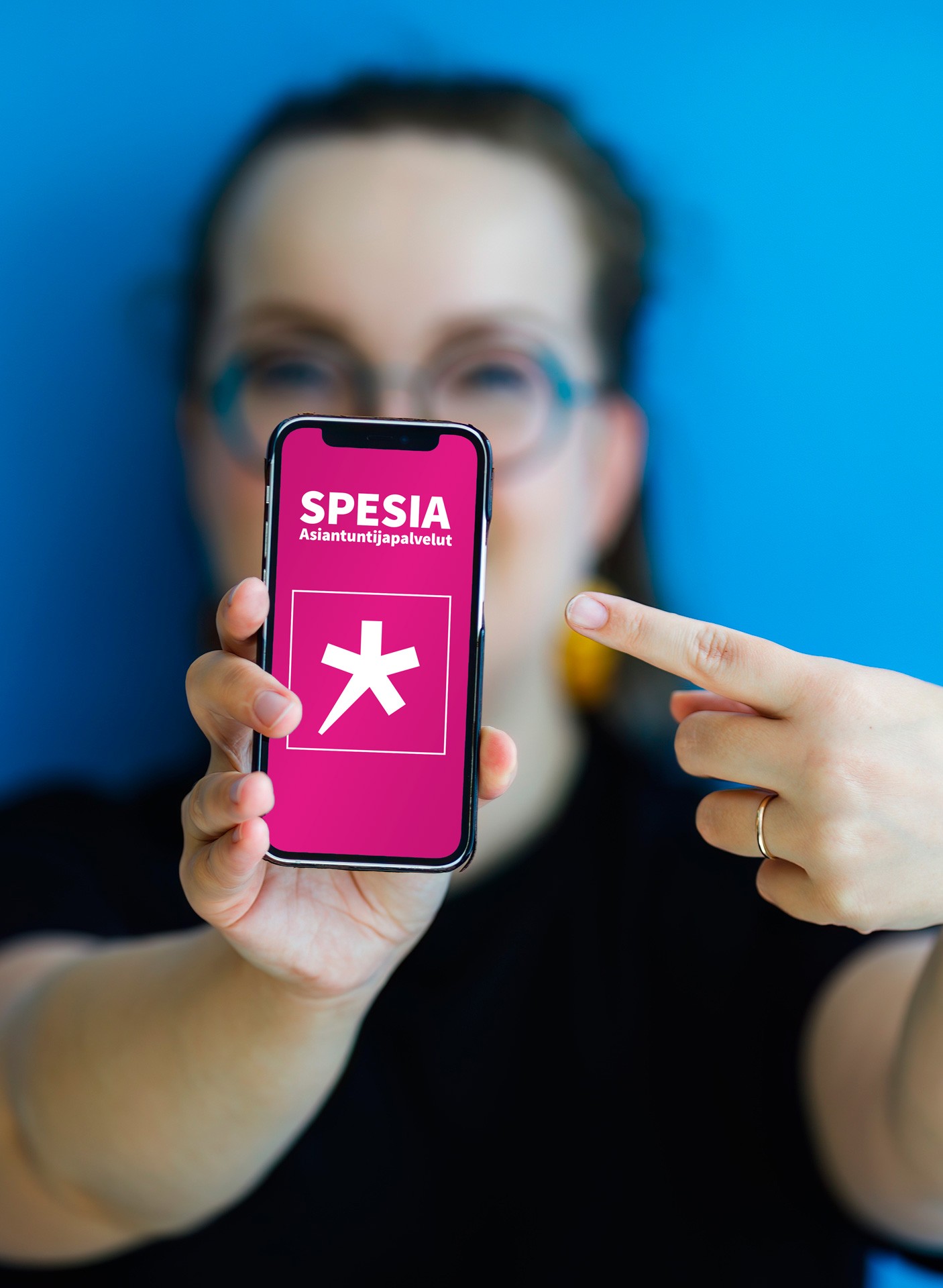 Nainen ojentaa kännykkää eteensä ja näyttää sormella ruudulla pinkillä pohjalla olevaa Spesia Asiantuntijapalveluiden logoa.