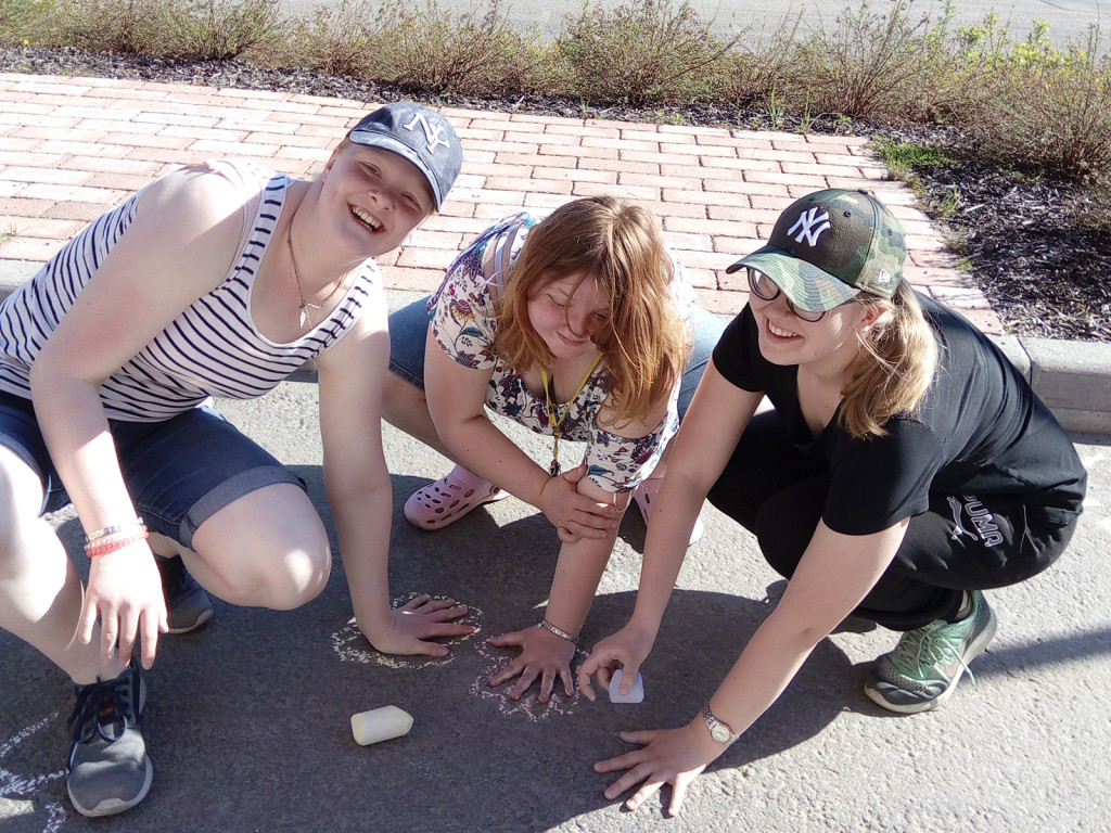Kolme nauravaista nuorta naista on kyykyllään ja pitää käsiään asfalttiin piirrettyjen kädenkuvien päällä.