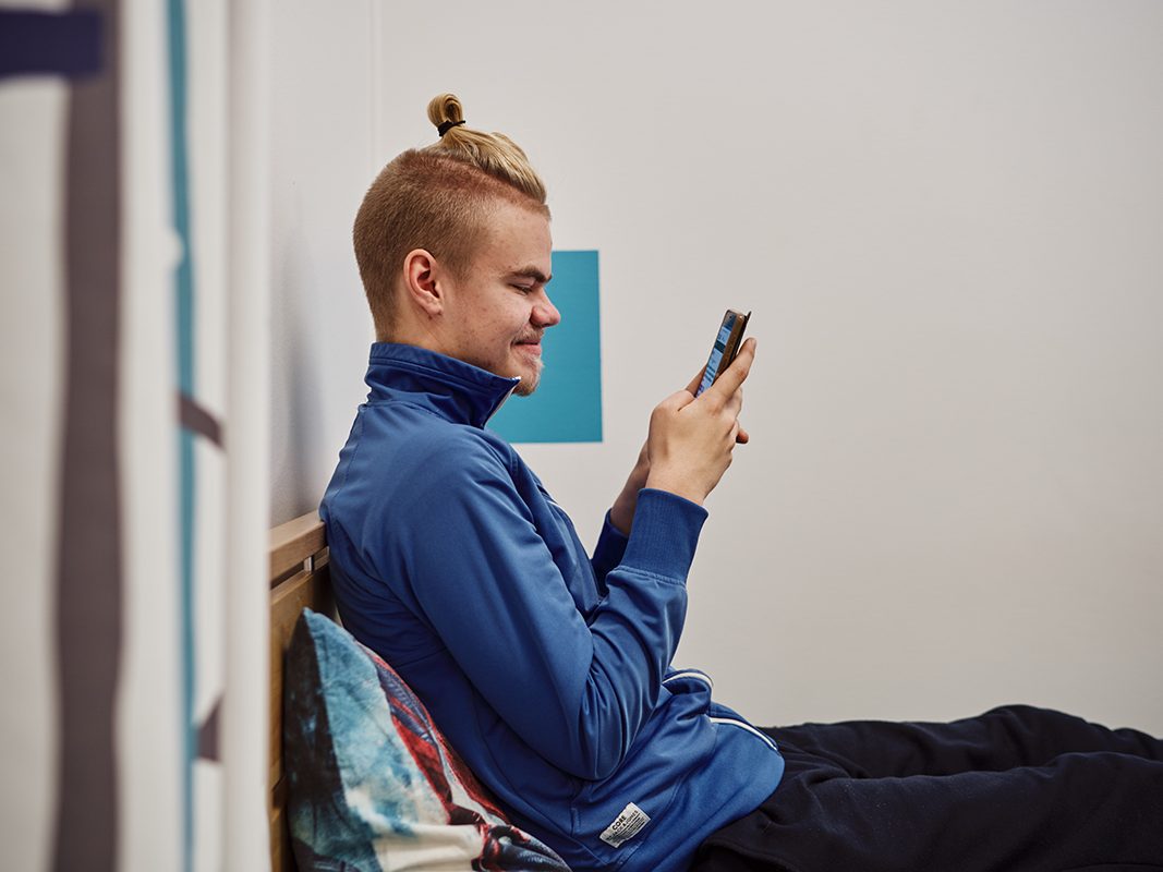 Nuori mies istuu sängyllä ja katsoo kännykkää.