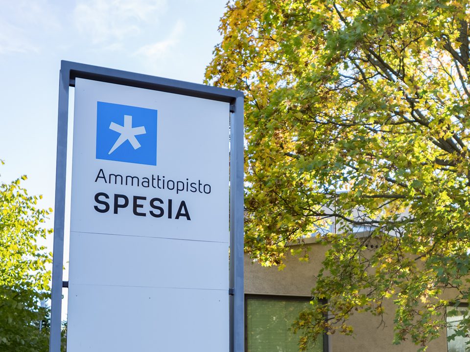 Ammattiopisto spesian kyltti auringossa loistavan syksyisen lehtipuun vieressä.