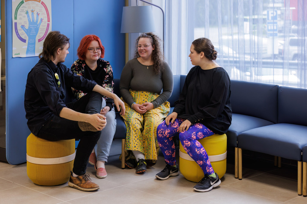 Neljä henkilöä istuu jakkaroilla puoliympyrän muodossa, taustalla sininen seinä ja ikkuna.