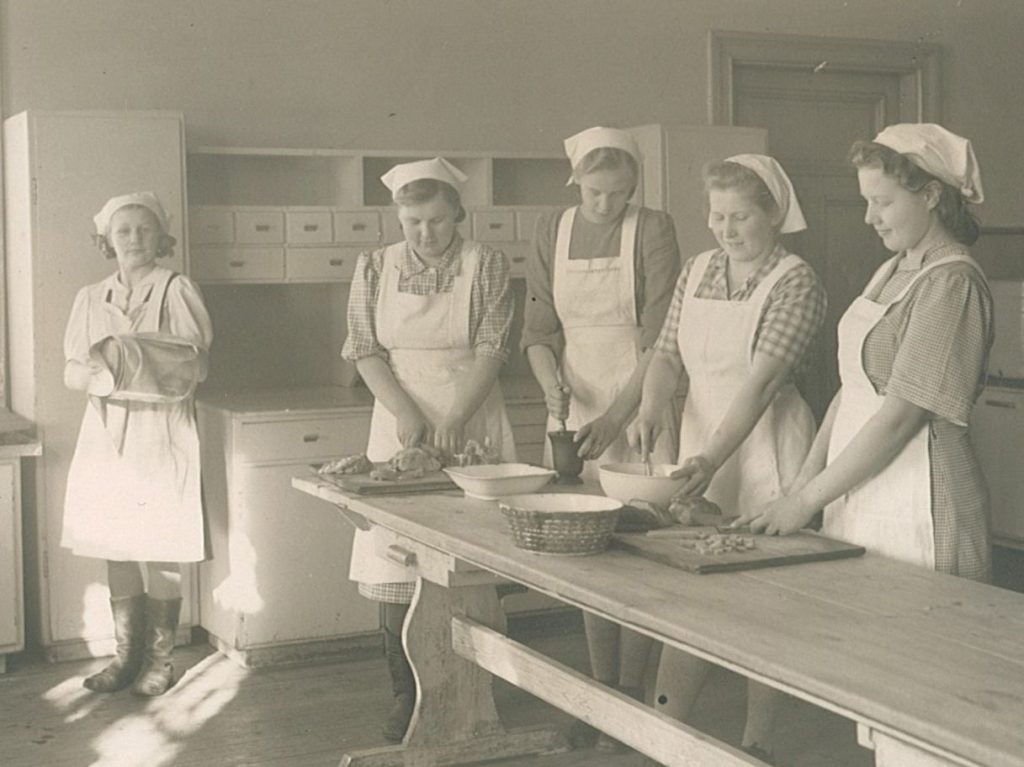 Valkoisiin essuihin ja päähineisiin pukeutuneet tytöt ja keittäjä tekevät ruokaa pirtinpöydän ääressä.